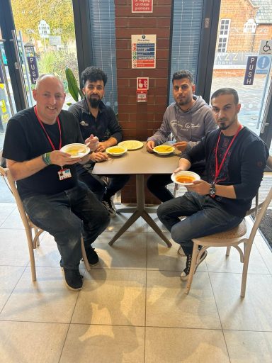 Four men sitting at a table holding bowls of food, smiling at the camera.