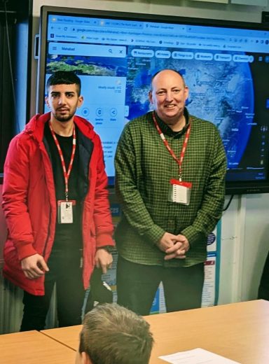 Two men stand in front of a large screen displaying a map, smiling and engaged.