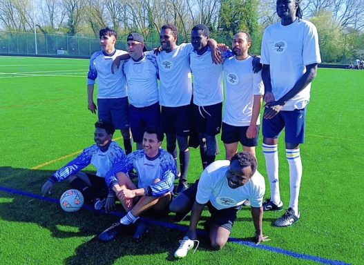 A group of eight male football players posing on a pitch, smiling and wearing team jerseys.